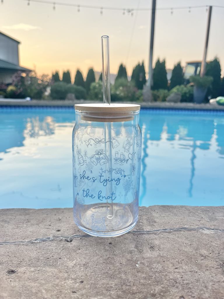 Clear glass cup with bamboo lid and straw featuring blue “She’s Tying the Knot” text and wedding doodles, perfect for brides and iced coffee lovers.