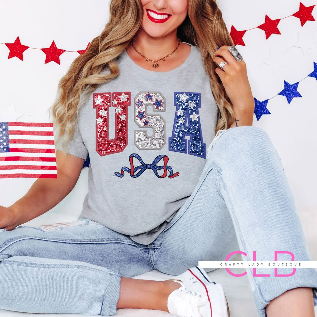"Woman wearing a light gray USA sequin t-shirt with red, silver, and blue lettering, sitting on the floor with a mini American flag, surrounded by patriotic star decorations."