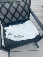 White Sarong with 'Mrs. Robertson' printed on it, draped over a chair on a wooden deck.