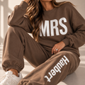 A woman wearing a matching brown sweatshirt and jogger pants set with white lettering, sitting in a sunlit indoor setting. The sweatshirt reads 'MRS' and the joggers read 'Haubert'.