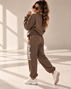 A woman modeling a matching brown sweatshirt and jogger sweatpants set. The joggers feature the brand name 'Haubert' printed vertically in white on the left leg.