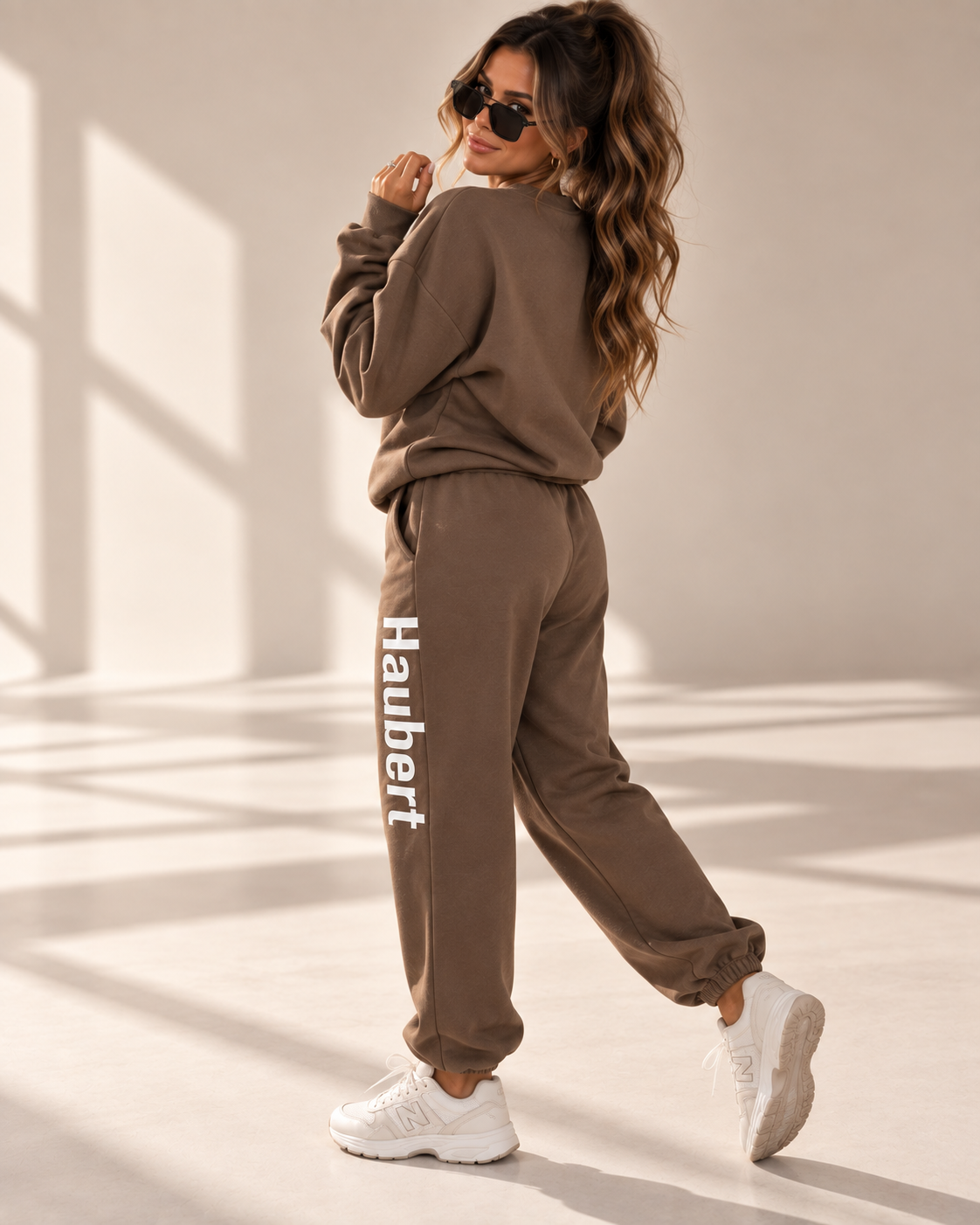 A woman modeling a matching brown sweatshirt and jogger sweatpants set. The joggers feature the brand name 'Haubert' printed vertically in white on the left leg.
