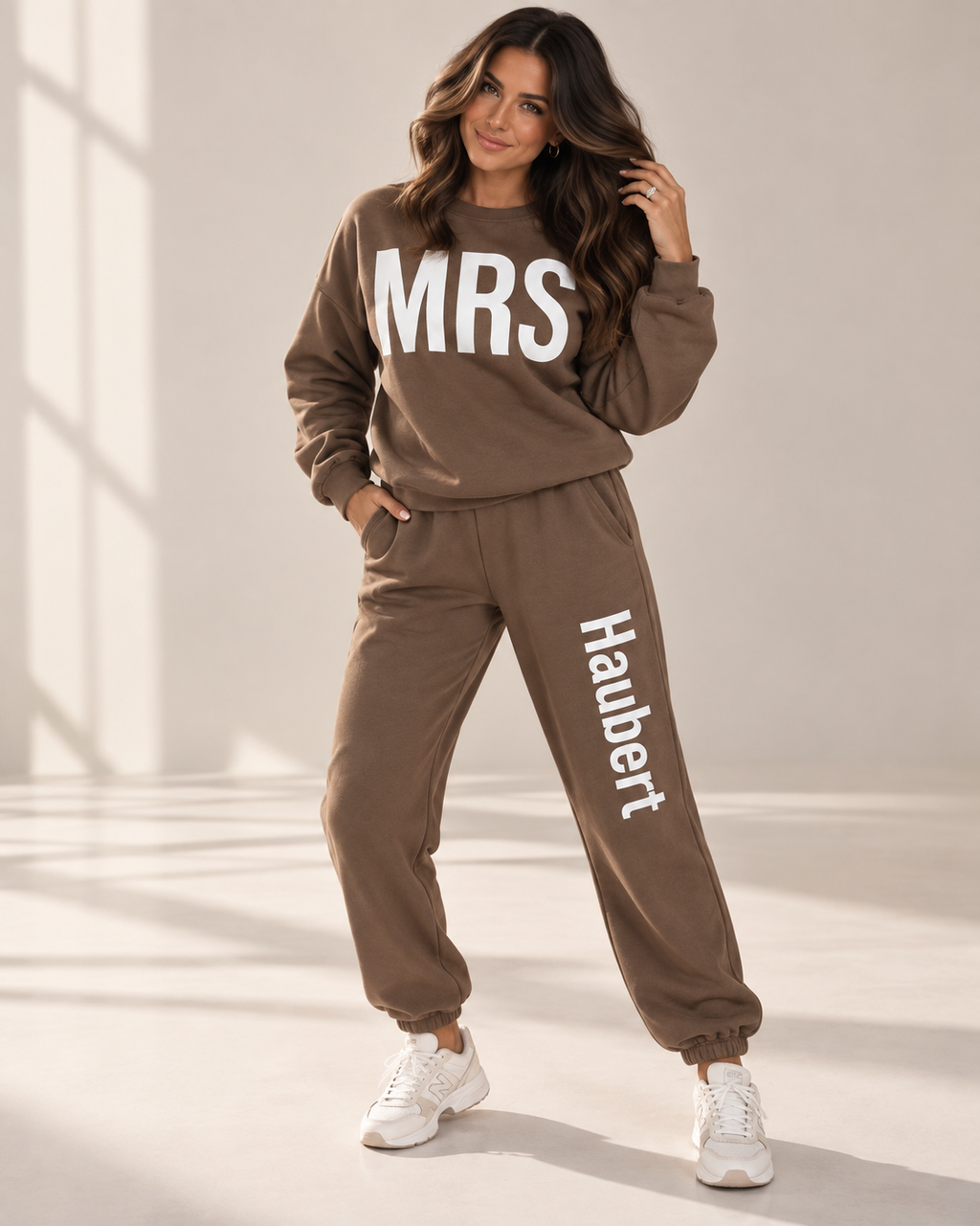A woman modeling a matching brown sweatshirt and jogger pants set with white text reading 'MRS' on the chest and 'Haubert' down the leg, paired with white sneakers in a brightly lit studio setting.
