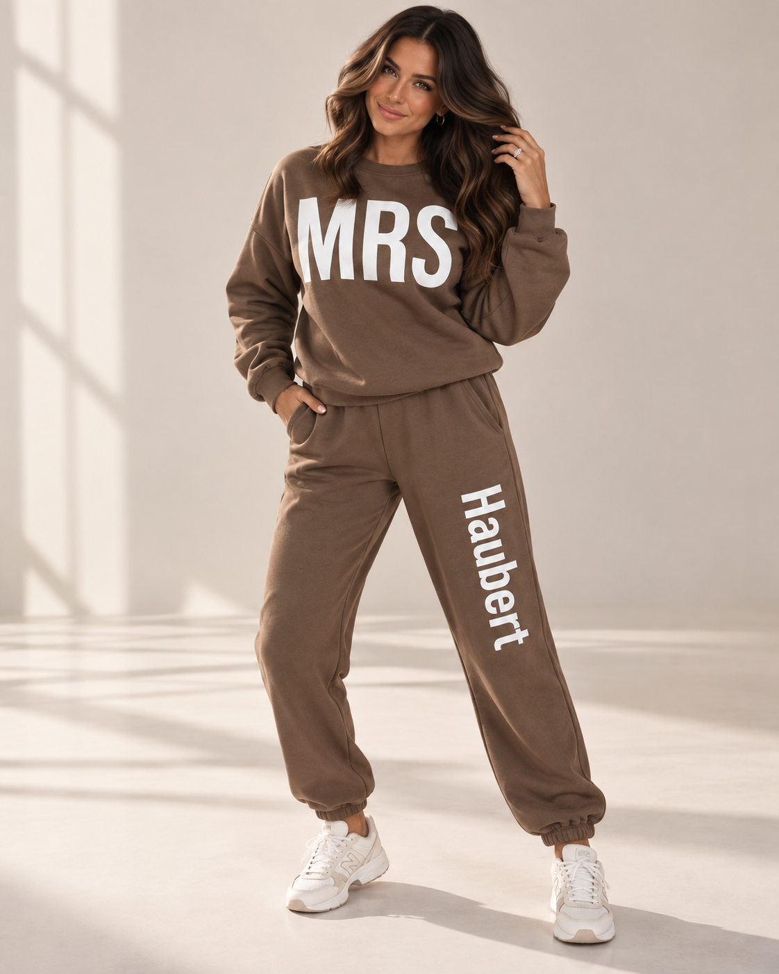 A woman modeling a matching brown sweatshirt and jogger pants set with white text reading 'MRS' on the chest and 'Haubert' down the leg, paired with white sneakers in a brightly lit studio setting.
