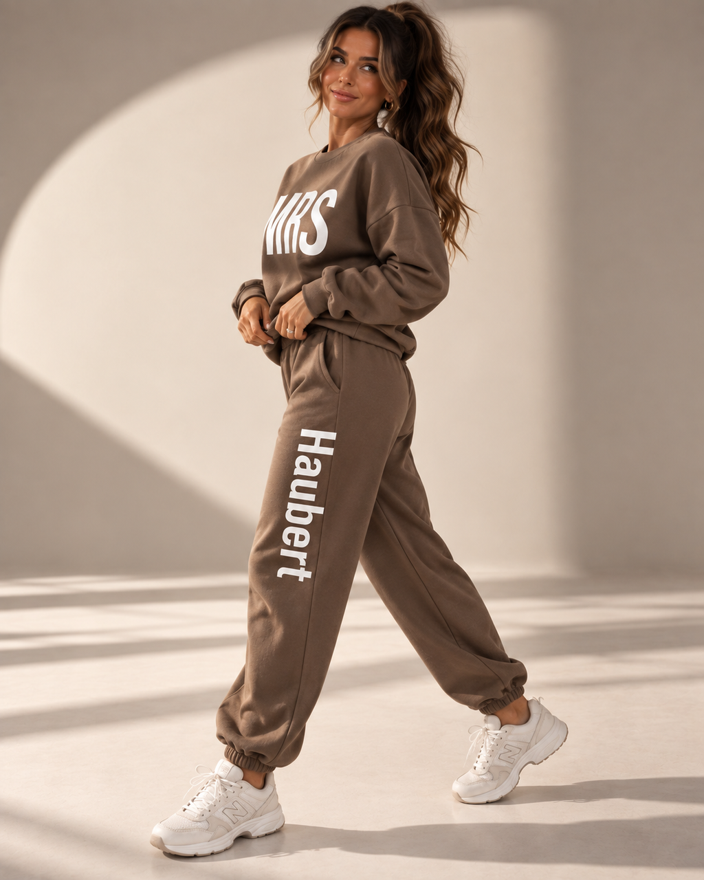 A woman modeling a matching brown sweatshirt and jogger pants set with white text branding, styled with white sneakers in a sunlit studio setting.