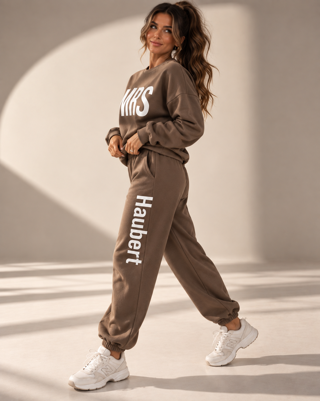 A woman modeling a matching brown sweatshirt and jogger pants set with white text branding, styled with white sneakers in a sunlit studio setting.