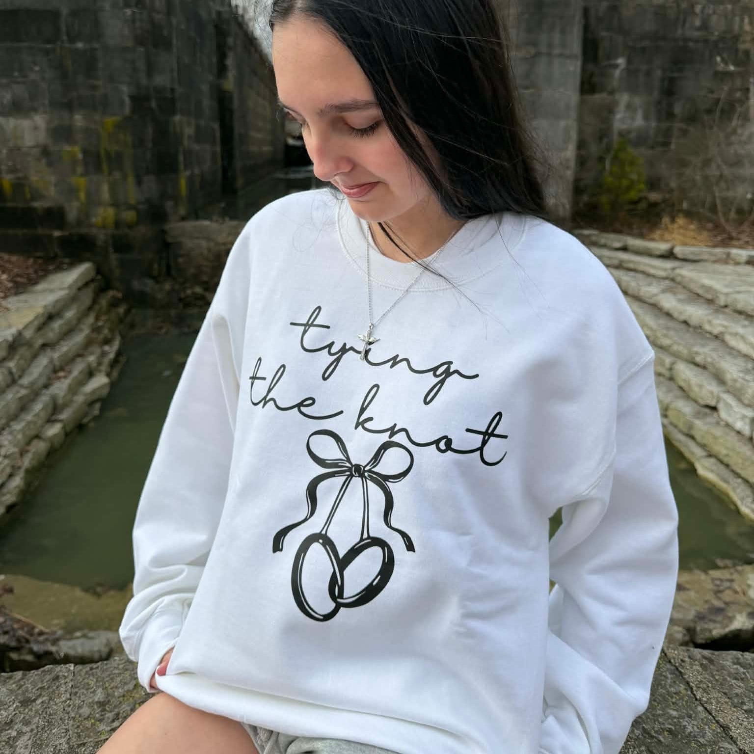 White bridal sweatshirt with “Tying the Knot” text and wedding ring bow design worn by engaged woman outdoors.
